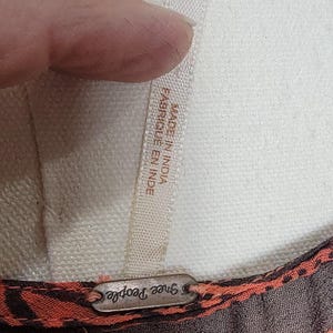 Puede incluir: Primer plano de una etiqueta de prenda con el texto "XS TP" y "MADE IN INDIA FABRIQUE EN INDE". La prenda tiene un dise&ntilde;o estampado marr&oacute;n y naranja. La marca "Free People" es visible.