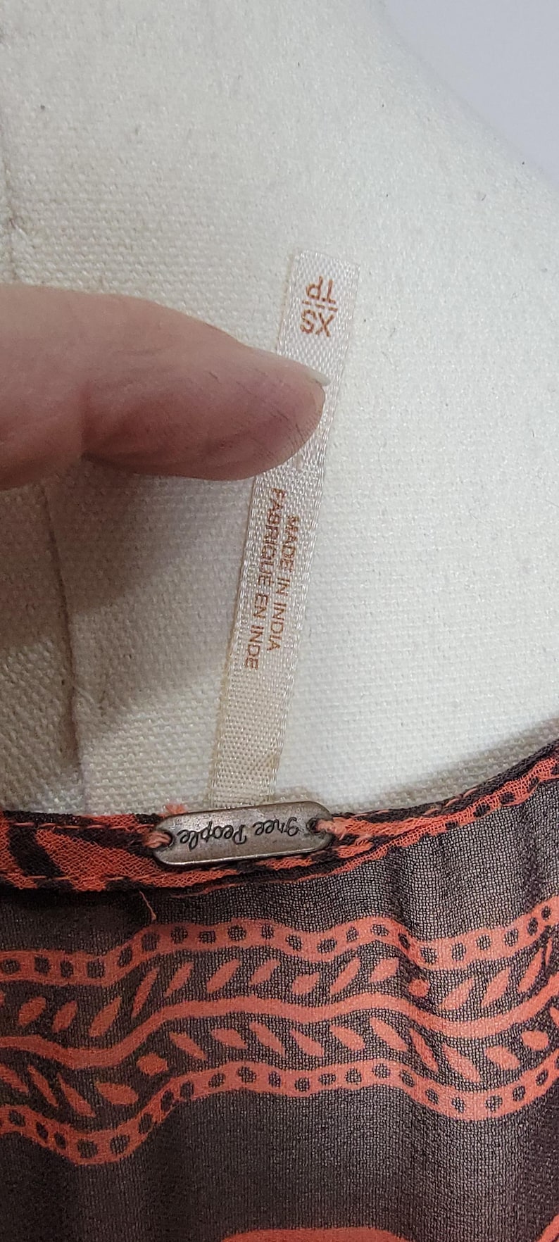 Puede incluir: Primer plano de una etiqueta de prenda con el texto "XS TP" y "MADE IN INDIA FABRIQUE EN INDE". La prenda tiene un dise&ntilde;o estampado marr&oacute;n y naranja. La marca "Free People" es visible.