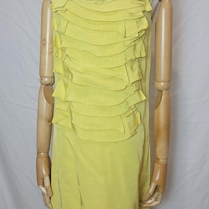 May include: A sleeveless, yellow dress with a layered ruffle design on the bodice. A small, black accent is placed at the neckline. The dress is displayed on a wooden mannequin.