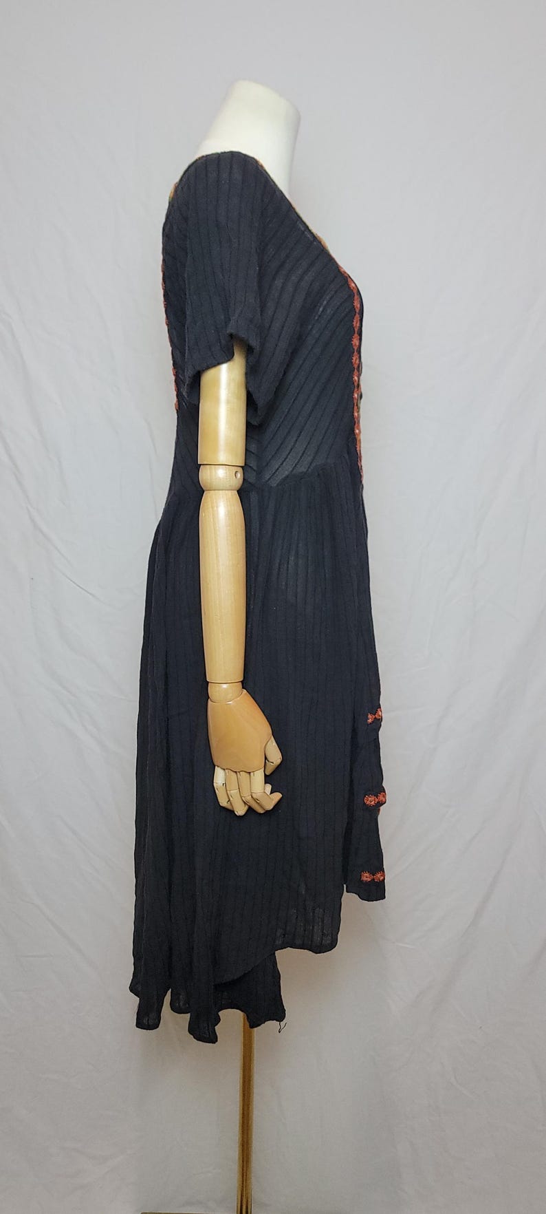 Puede incluir: Vestido negro de manga corta con textura acanalada vertical. El vestido presenta bordados naranjas a lo largo del escote y en el lateral. El vestido se exhibe en un maniqu&iacute;.