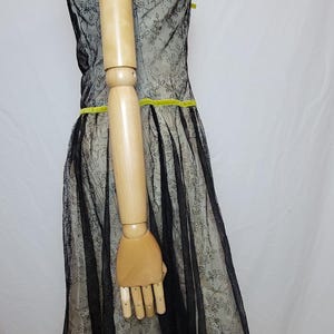 Pu&ograve; includere: Un abito in stile vintage con un rivestimento in pizzo nero su un tessuto di colore chiaro. L'abito presenta un corpetto aderente, una gonna svasata e una cintura in nastro giallo brillante. L'abito &egrave; esposto su un manichino.