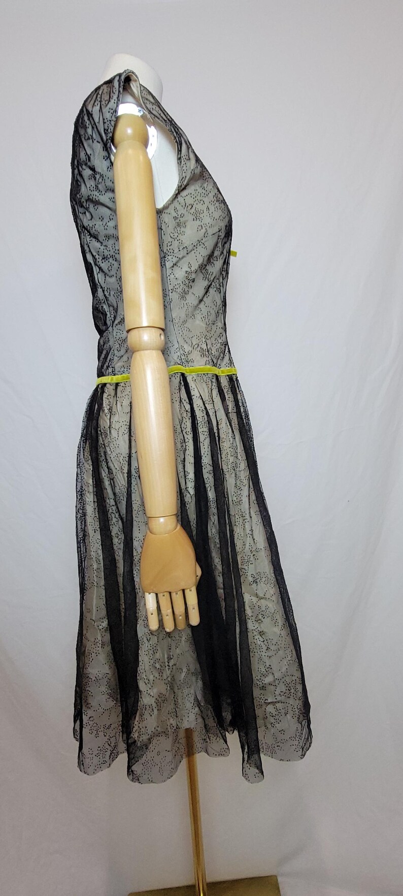 Pu&ograve; includere: Un abito in stile vintage con un rivestimento in pizzo nero su un tessuto di colore chiaro. L'abito presenta un corpetto aderente, una gonna svasata e una cintura in nastro giallo brillante. L'abito &egrave; esposto su un manichino.
