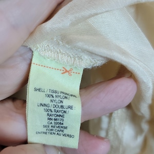 Pu&ograve; includere: Primo piano di un'etichetta di tessuto giallo chiaro con testo in inglese e francese. L'etichetta recita "SHELL/TISSU PRINCIPAL: 100% NYLON/NYLON" e "LINING/DOUBLURE: 100% RAYON/RAYONNE".