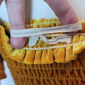 May include: Close-up of a mustard yellow velvet garment with a gathered texture. A white tag with text is held up, reading "S-P Made In USA of imported fabric/Fabriqu&eacute; Aux E-U. du". The fabric appears soft and luxurious.