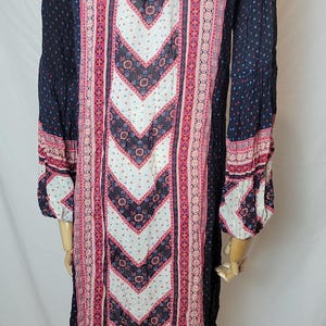 May include: A long-sleeved dress with a navy blue background and a central white chevron pattern. The dress features pink and red floral and geometric designs, with matching patterns on the sleeves. The dress is made of a lightweight fabric.