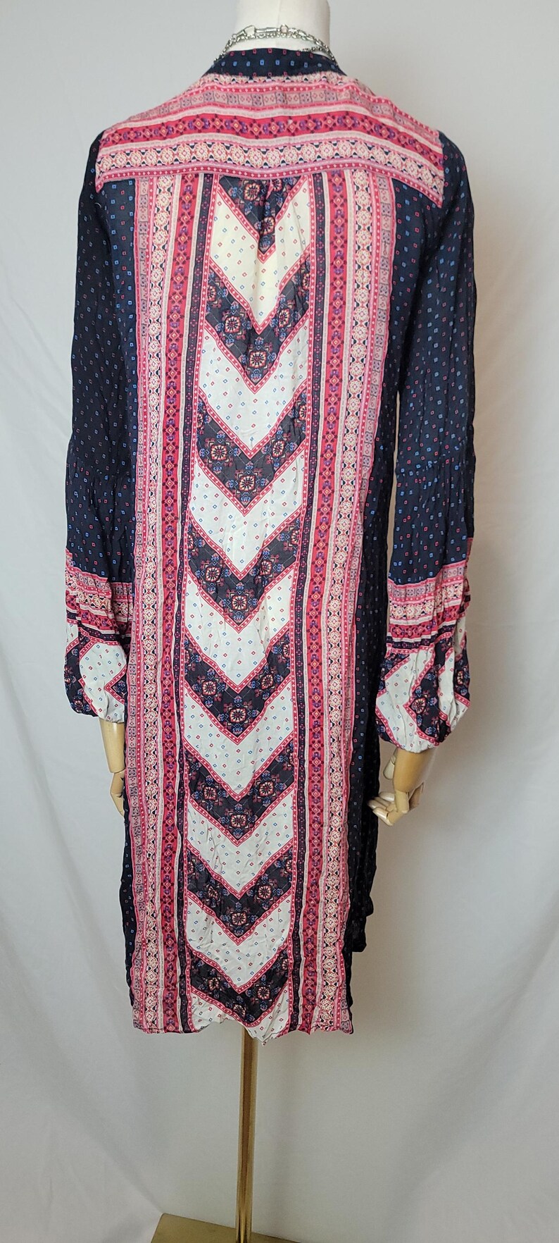 May include: A long-sleeved dress with a navy blue background and a central white chevron pattern. The dress features pink and red floral and geometric designs, with matching patterns on the sleeves. The dress is made of a lightweight fabric.