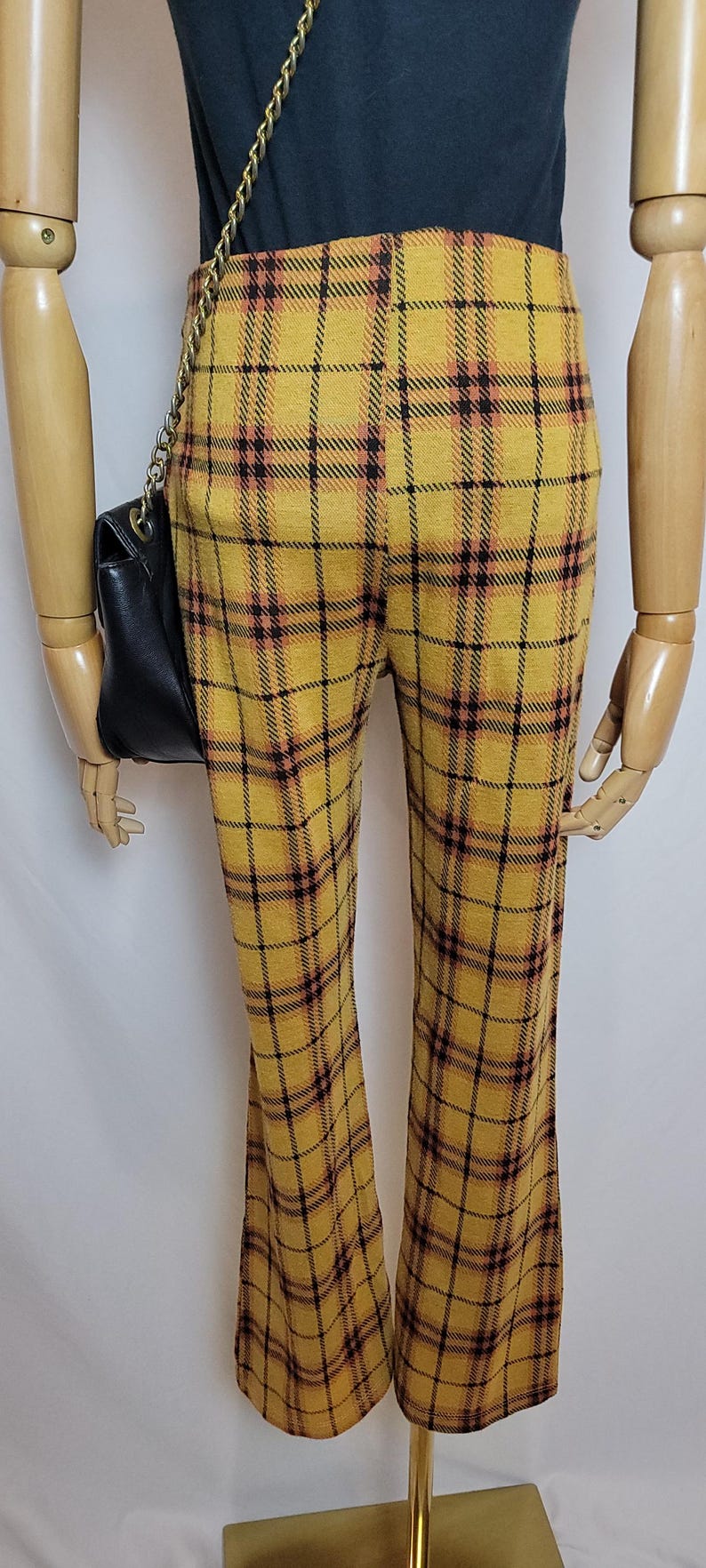 K&ouml;nnte beinhalten: Senfgelbe karierte Hose mit einem schwarz-roten Gittermuster. Die Hose hat ein ausgestelltes Bein und wird auf einer Schaufensterpuppe gezeigt. Eine schwarze Handtasche mit goldenem Kettentr&auml;ger ist sichtbar.