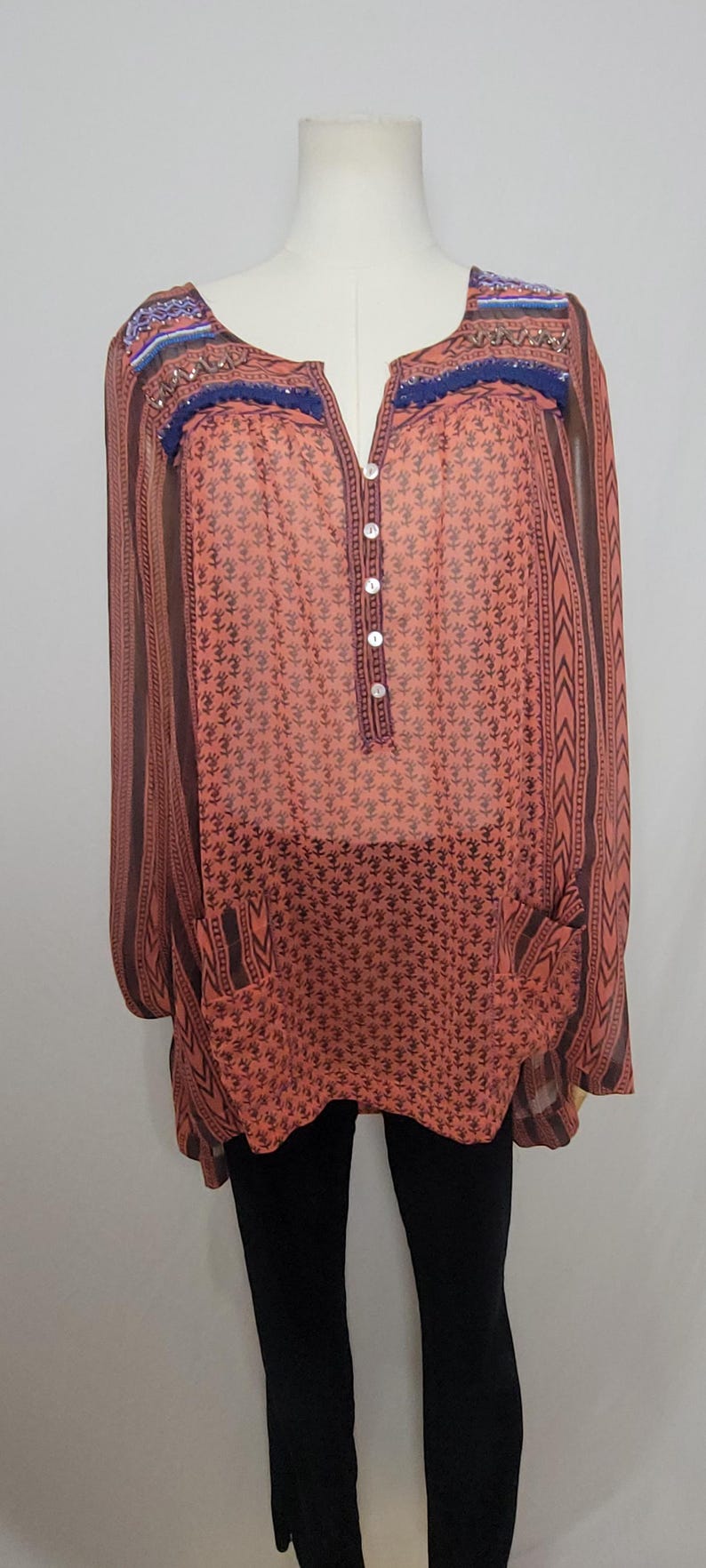 Puede incluir: T&uacute;nica color coral con estampado floral y mangas largas. La parte superior presenta una botonadura frontal, dos bolsillos y adornos decorativos a lo largo del escote. Las mangas y los laterales tienen un dise&ntilde;o a rayas. La t&uacute;nica se exhibe en un maniqu&iacute;.
