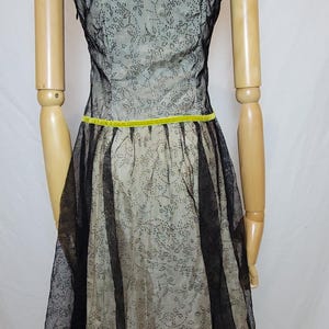 Pu&ograve; includere: Abito vintage su manichino. L'abito presenta un rivestimento in pizzo floreale nero su un tessuto colorato chiaro. Un nastro giallo brillante accentua la vita. L'abito ha maniche corte e una gonna svasata.