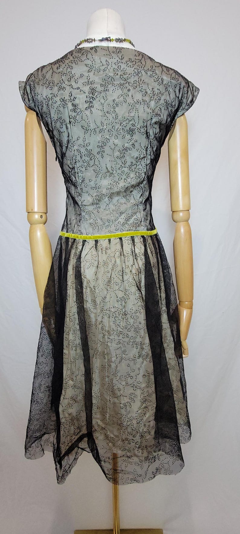 Pu&ograve; includere: Abito vintage su manichino. L'abito presenta un rivestimento in pizzo floreale nero su un tessuto colorato chiaro. Un nastro giallo brillante accentua la vita. L'abito ha maniche corte e una gonna svasata.