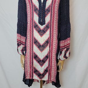 May include: A long-sleeved tunic dress with a V-neck, displayed on a mannequin. The dress features a navy blue background with a patterned design in pink, white, and red. The sleeves have a dotted pattern.