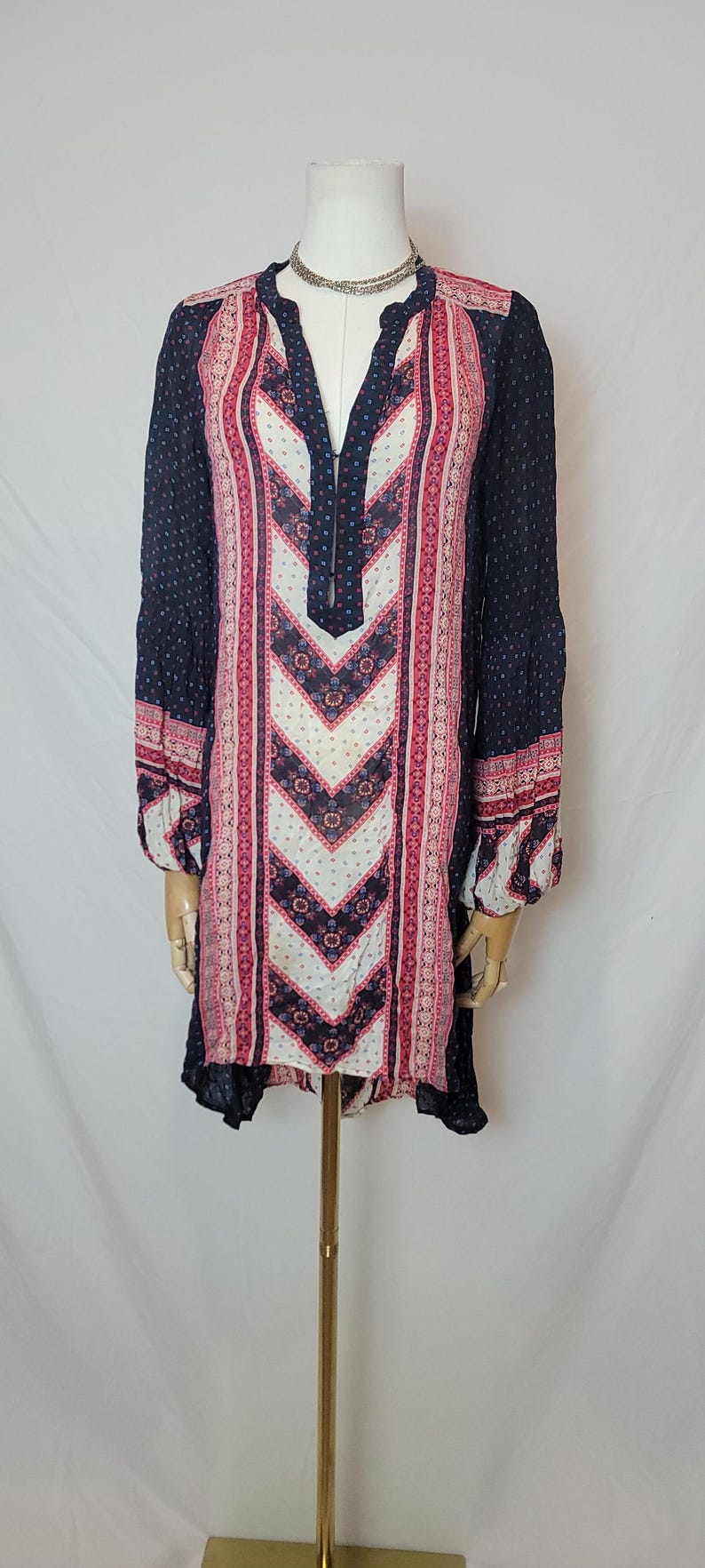 May include: A long-sleeved tunic dress with a V-neck, displayed on a mannequin. The dress features a navy blue background with a patterned design in pink, white, and red. The sleeves have a dotted pattern.