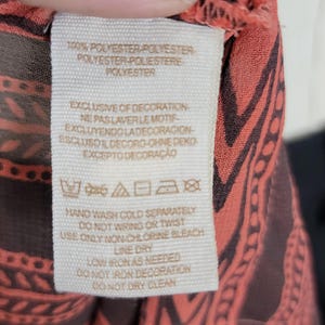 Puede incluir: Primer plano de una etiqueta de ropa con instrucciones de cuidado y composici&oacute;n de la tela. La etiqueta es de color crema con texto negro. La tela tiene un dise&ntilde;o coral y marr&oacute;n.