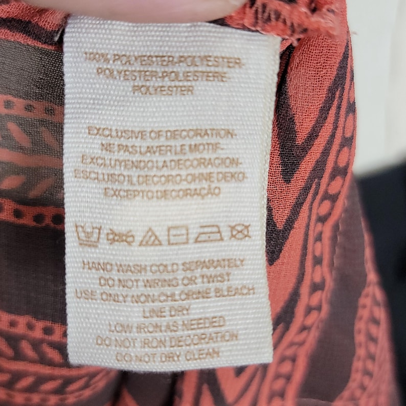Puede incluir: Primer plano de una etiqueta de ropa con instrucciones de cuidado y composici&oacute;n de la tela. La etiqueta es de color crema con texto negro. La tela tiene un dise&ntilde;o coral y marr&oacute;n.