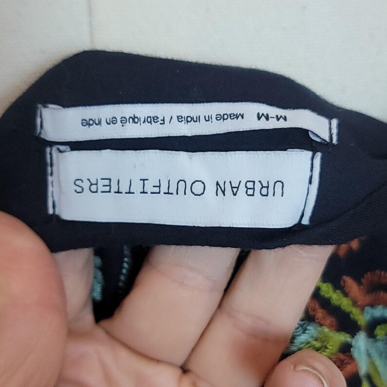 May include: Black garment with a white tag that reads "URBAN OUTFITTERS" in capital letters. Below the tag is another tag that reads "Made in India / Fabriqu&eacute; en Inde". The garment has colourful embroidery.