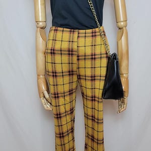 K&ouml;nnte beinhalten: Eine Schaufensterpuppe tr&auml;gt ein dunkelblaues T-Shirt, eine gelbe karierte Hose und eine schwarze Handtasche mit goldenem Kettenriemen. Die Hose hat ein ausgestelltes Bein und ein gelbes, schwarzes und rotes Karomuster. Die Handtasche h&auml;ngt an der Seite.