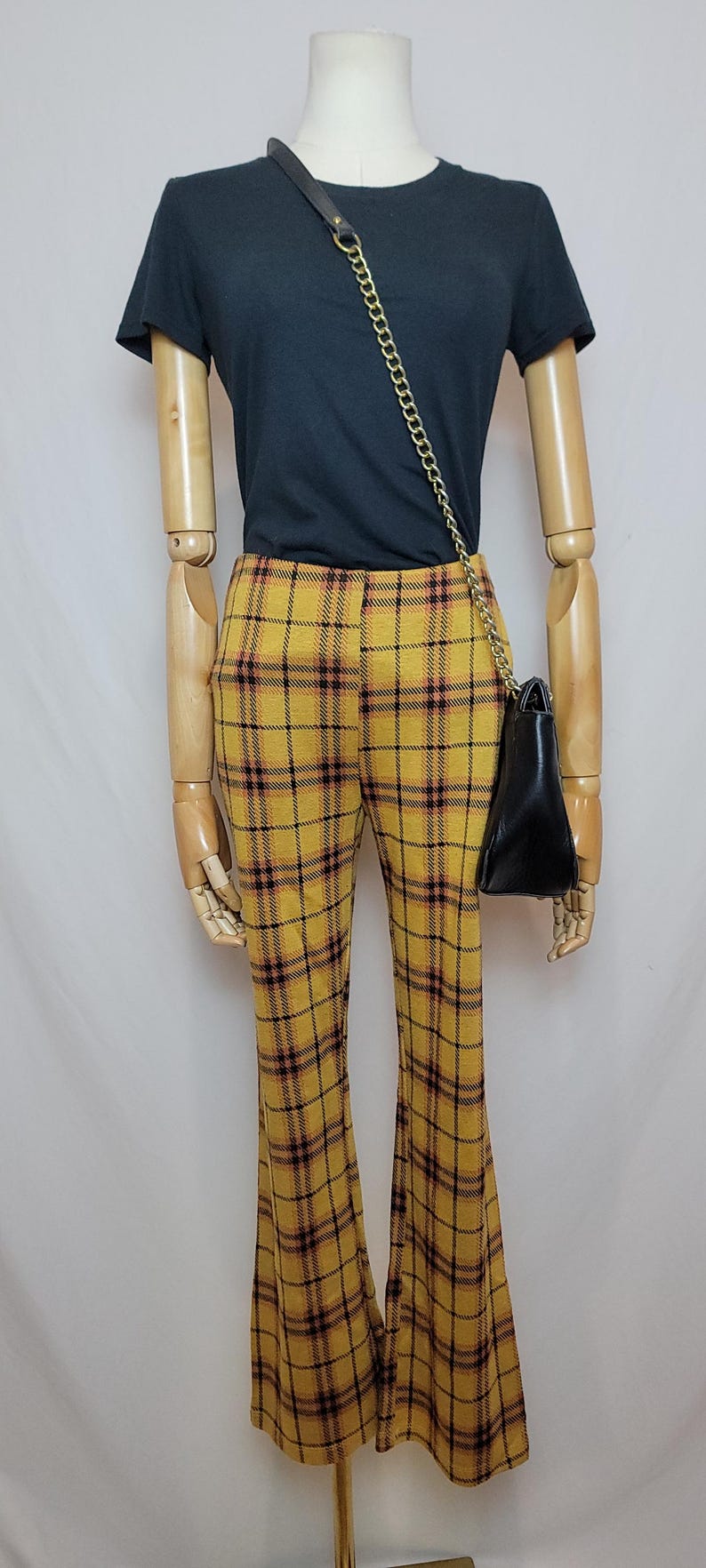K&ouml;nnte beinhalten: Eine Schaufensterpuppe tr&auml;gt ein dunkelblaues T-Shirt, eine gelbe karierte Hose und eine schwarze Handtasche mit goldenem Kettenriemen. Die Hose hat ein ausgestelltes Bein und ein gelbes, schwarzes und rotes Karomuster. Die Handtasche h&auml;ngt an der Seite.
