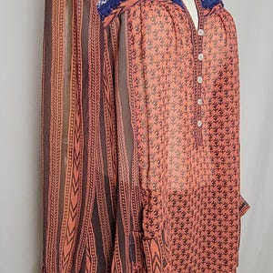 Puede incluir: Blusa color coral con estampado floral y mangas a rayas marrones. El escote y los hombros est&aacute;n adornados con adornos de cuentas azules y naranjas. La blusa tiene una parte delantera abotonada y mangas largas.