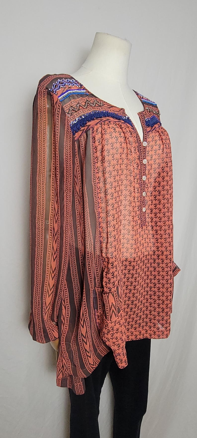 Puede incluir: Blusa color coral con estampado floral y mangas a rayas marrones. El escote y los hombros est&aacute;n adornados con adornos de cuentas azules y naranjas. La blusa tiene una parte delantera abotonada y mangas largas.