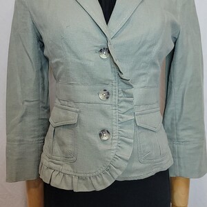 May include: A pale green blazer with a ruffled front and three buttons. The blazer features two front pockets and three-quarter sleeves. The garment is displayed on a mannequin.