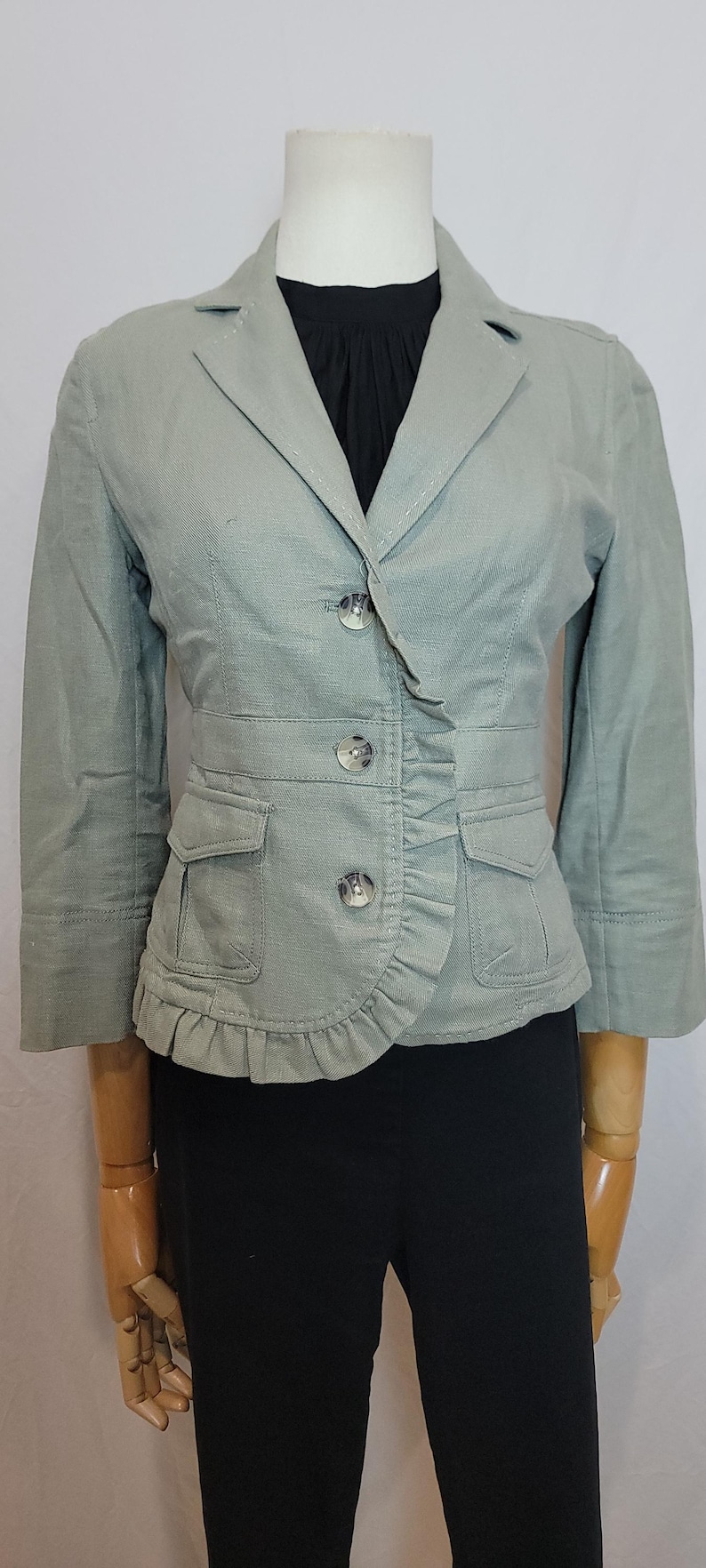 May include: A pale green blazer with a ruffled front and three buttons. The blazer features two front pockets and three-quarter sleeves. The garment is displayed on a mannequin.