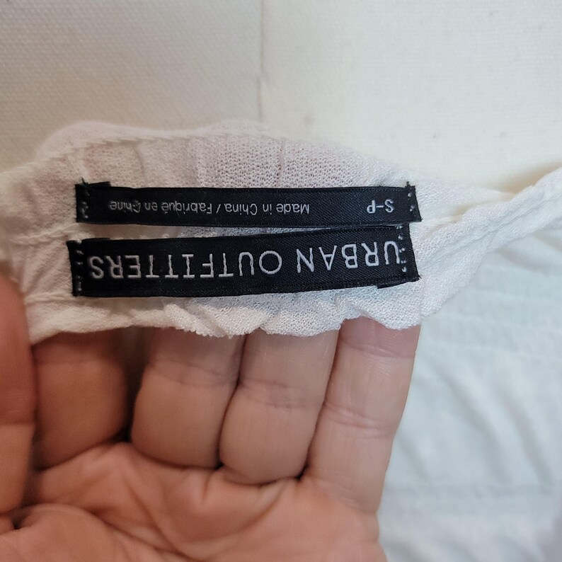 Op de afbeelding: Close-up van een wit kledinglabel met de tekst "URBAN OUTFITTERS" in wit op een zwarte achtergrond. Onder het label staat een ander label met de tekst "Made in China / Fabriqu&eacute; en Chine" en "S-P".