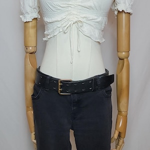 Op de afbeelding: Een witte, geplooid crop top met een trekkoord detail wordt gecombineerd met een zwarte jeans en een zwarte riem met een gouden gesp. Een zilveren ketting wordt om de nek van de mannequin gedragen.