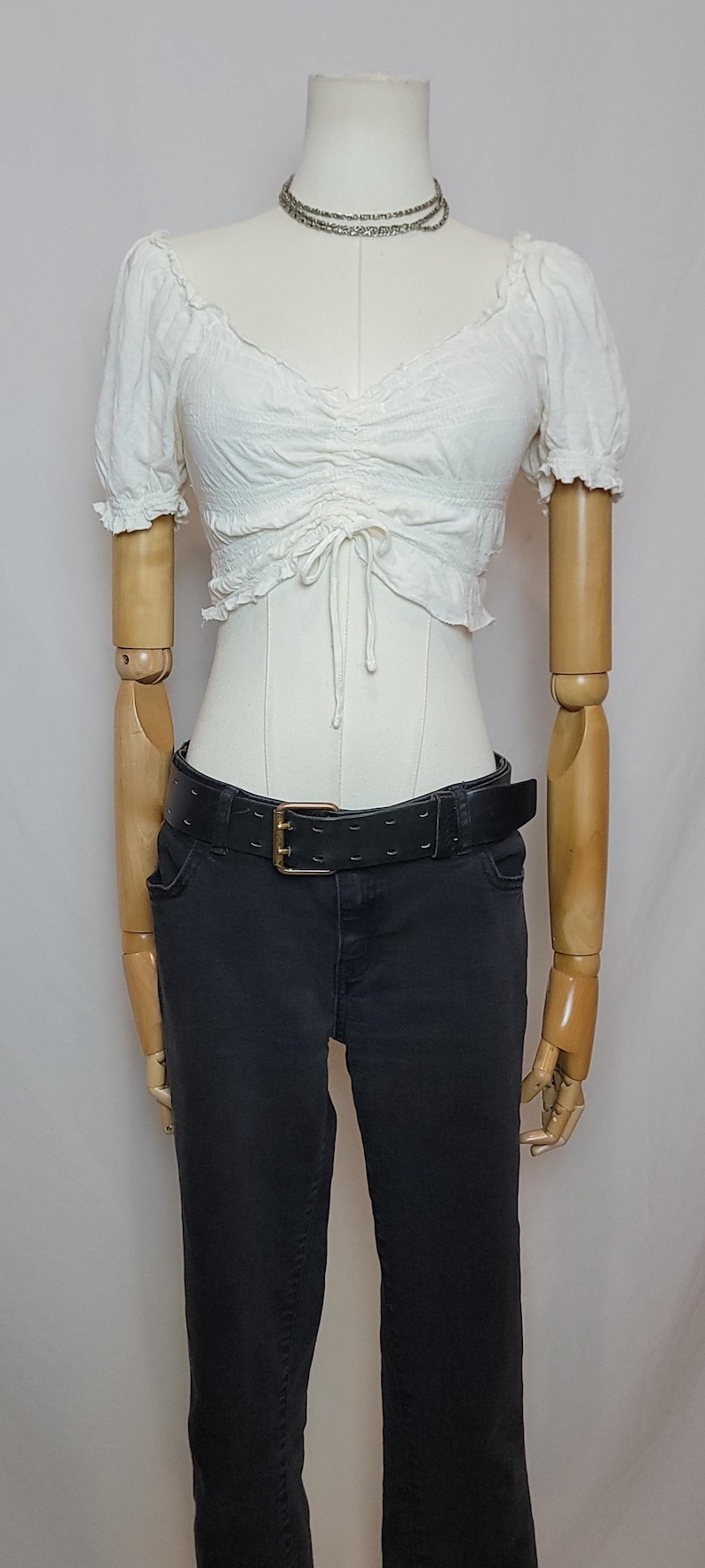 Op de afbeelding: Een witte, geplooid crop top met een trekkoord detail wordt gecombineerd met een zwarte jeans en een zwarte riem met een gouden gesp. Een zilveren ketting wordt om de nek van de mannequin gedragen.