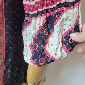 May include: A long-sleeved garment with a patterned design. The garment features a black background with small blue and red squares, and patterned sleeves with red, white, and black bands. The sleeve is gathered at the wrist.
