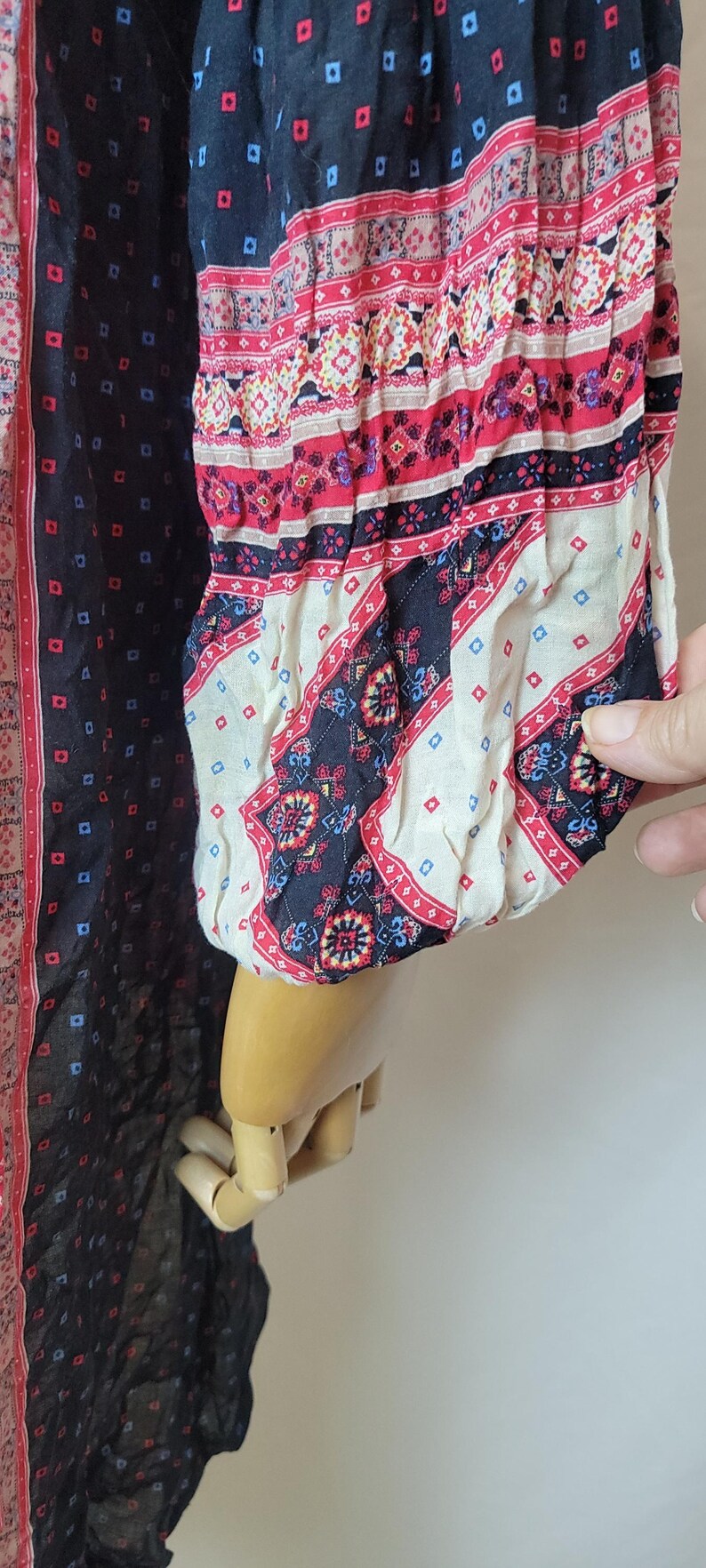 May include: A long-sleeved garment with a patterned design. The garment features a black background with small blue and red squares, and patterned sleeves with red, white, and black bands. The sleeve is gathered at the wrist.