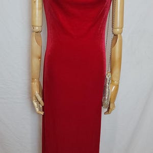 May include: A floor-length, red velvet dress with a cowl neckline. The dress is displayed on a mannequin with wooden arms. A small, silver beaded purse hangs from the mannequin's arm.