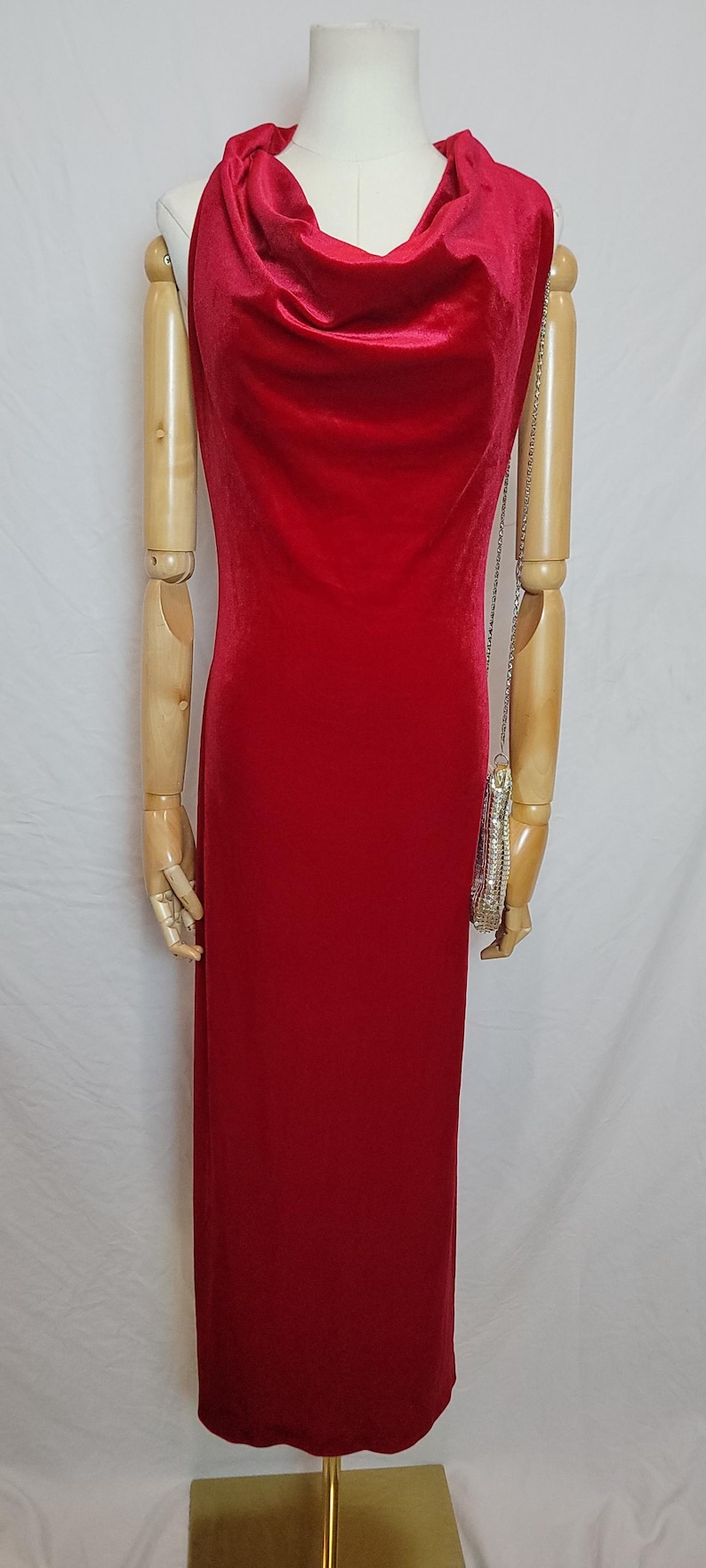 May include: A floor-length, red velvet dress with a cowl neckline. The dress is displayed on a mannequin with wooden arms. A small, silver beaded purse hangs from the mannequin's arm.