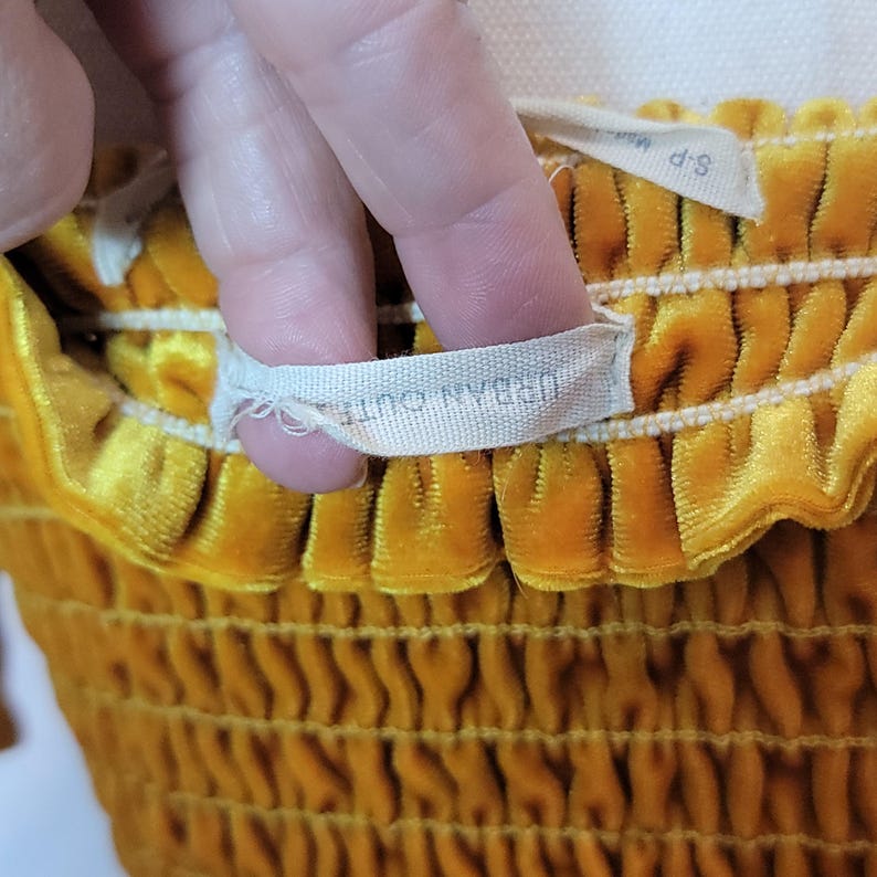 May include: Close-up of a mustard yellow velvet top with a smocked design. The tag reads "URBAN OUTFITTERS" and "S-P". The fabric has a soft, textured appearance.