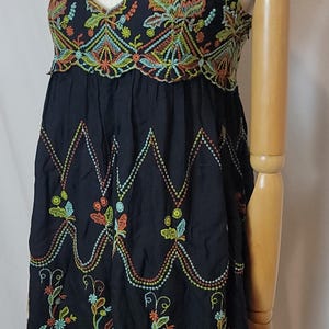 May include: Black dress with colourful floral embroidery. The dress has a V-neck and spaghetti straps. The embroidery includes orange, yellow, green, and blue details. The dress is displayed on a mannequin.