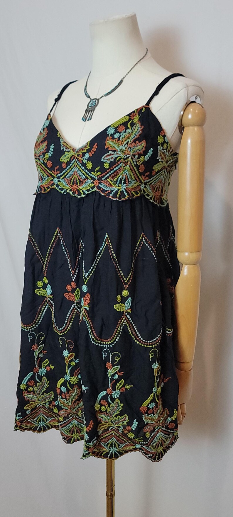 May include: Black dress with colourful floral embroidery. The dress has a V-neck and spaghetti straps. The embroidery includes orange, yellow, green, and blue details. The dress is displayed on a mannequin.