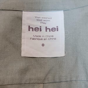 May include: A close-up shot of a pale green garment featuring a white label. The label displays the brand name "hei hei" in dark purple, with the phrases "Well washed, Well worn, Play" above. It also states "Made in China, Fabriqu&eacute; en Chine" and the number "0".