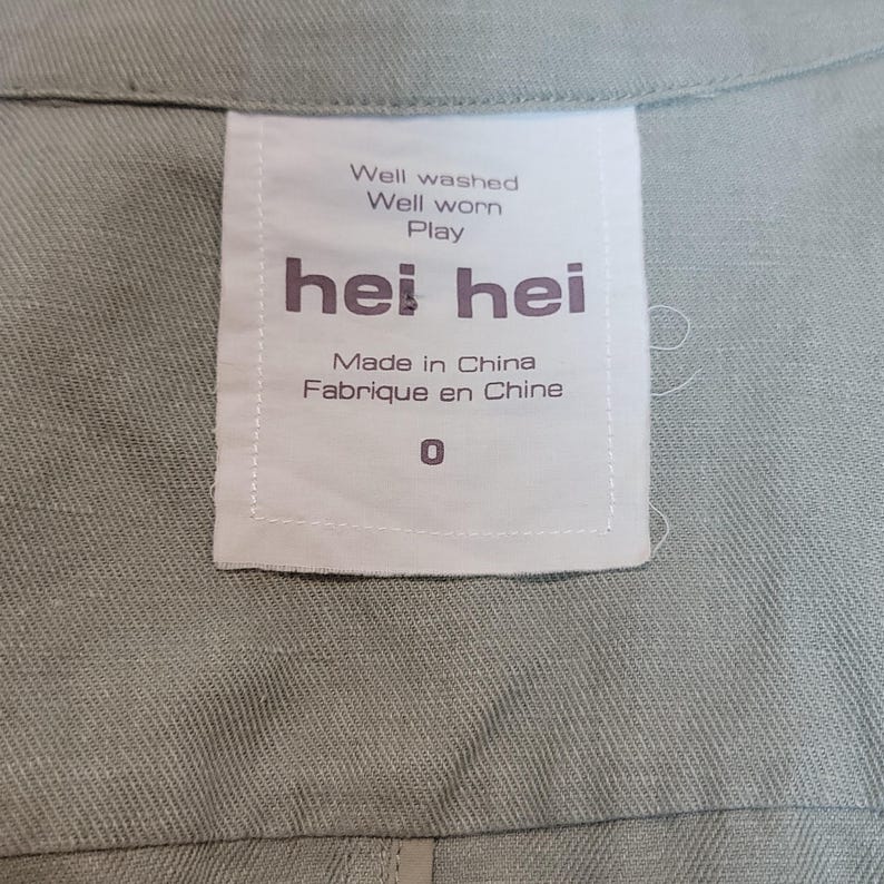 May include: A close-up shot of a pale green garment featuring a white label. The label displays the brand name "hei hei" in dark purple, with the phrases "Well washed, Well worn, Play" above. It also states "Made in China, Fabriqu&eacute; en Chine" and the number "0".