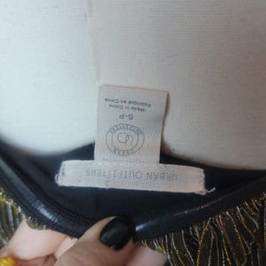 May include: Close-up of a black garment with gold fringe. The tag reads "Urban Outfitters" and "Made in China". The garment is size S-P.