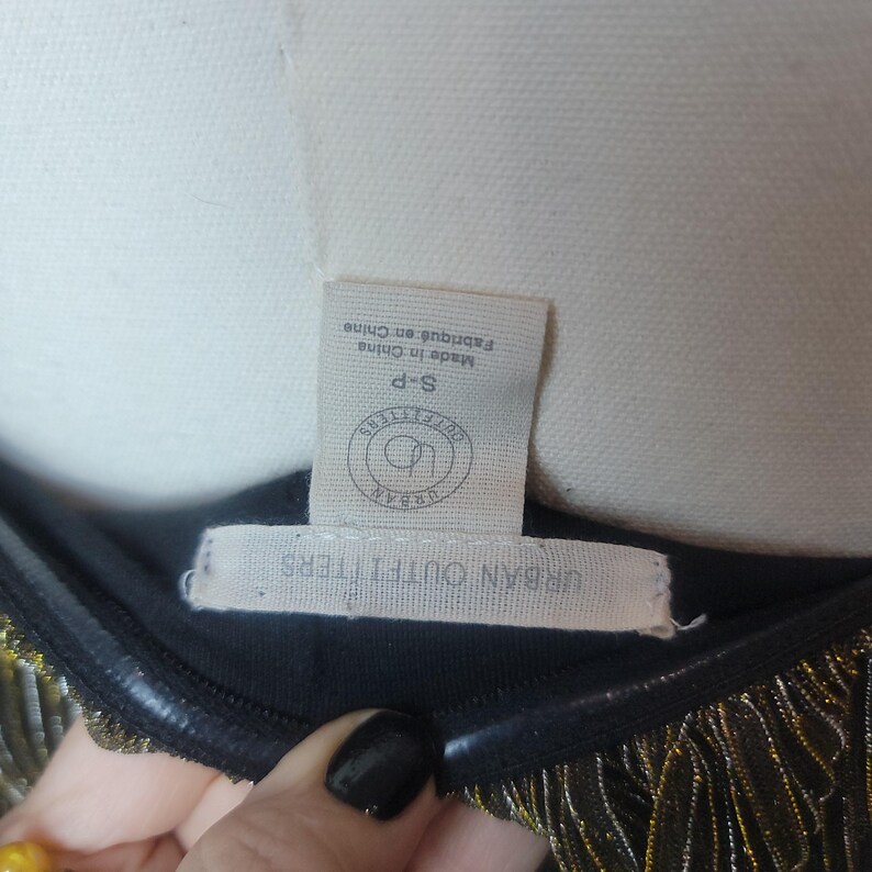 May include: Close-up of a black garment with gold fringe. The tag reads "Urban Outfitters" and "Made in China". The garment is size S-P.