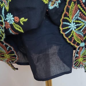May include: Close-up of a black garment with colourful floral embroidery. The embroidery features orange, green, and blue thread. The garment is displayed on a gold stand, showcasing the intricate details of the design.