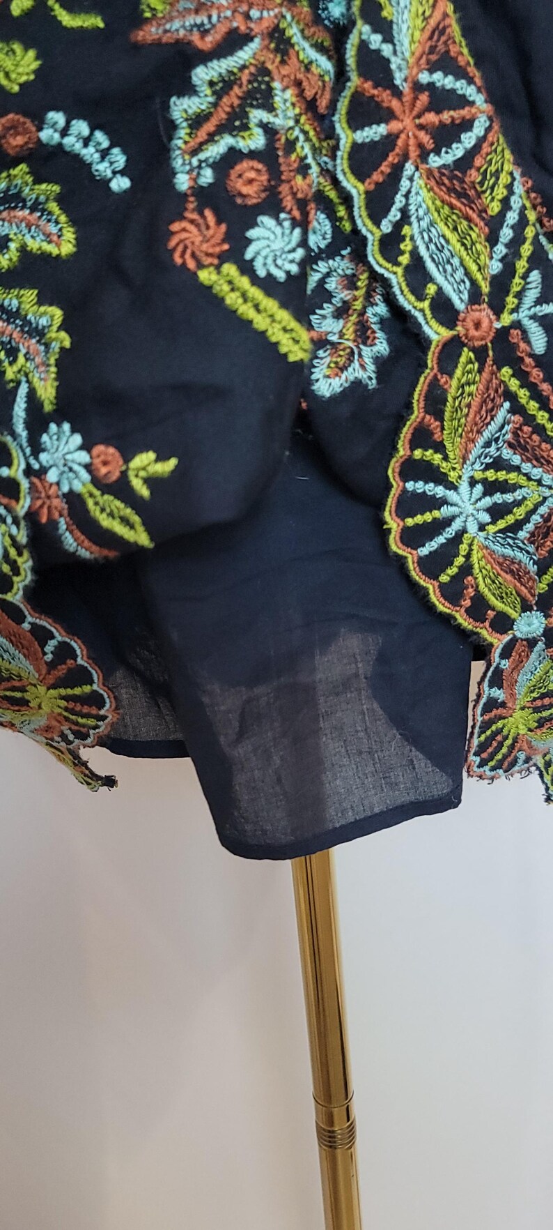 May include: Close-up of a black garment with colourful floral embroidery. The embroidery features orange, green, and blue thread. The garment is displayed on a gold stand, showcasing the intricate details of the design.