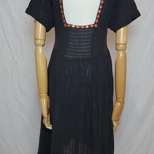 Puede incluir: Vestido negro con mangas cortas y escote cuadrado. El vestido presenta un ribete floral decorativo alrededor del escote y una espalda con abertura. El vestido se exhibe en un maniqu&iacute;.