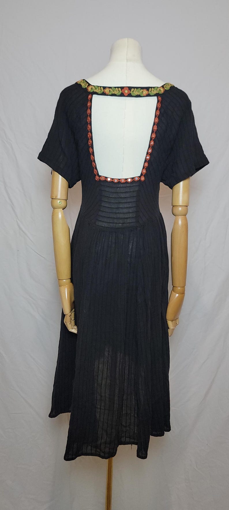 Puede incluir: Vestido negro con mangas cortas y escote cuadrado. El vestido presenta un ribete floral decorativo alrededor del escote y una espalda con abertura. El vestido se exhibe en un maniqu&iacute;.