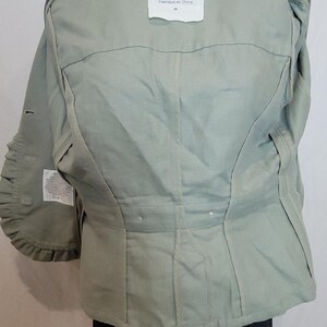 May include: A pale green jacket with a structured design. The jacket has a collar, pleats, and a label that reads "hei hei". The sleeves have a unique ruffled detail. The jacket is worn over black trousers.