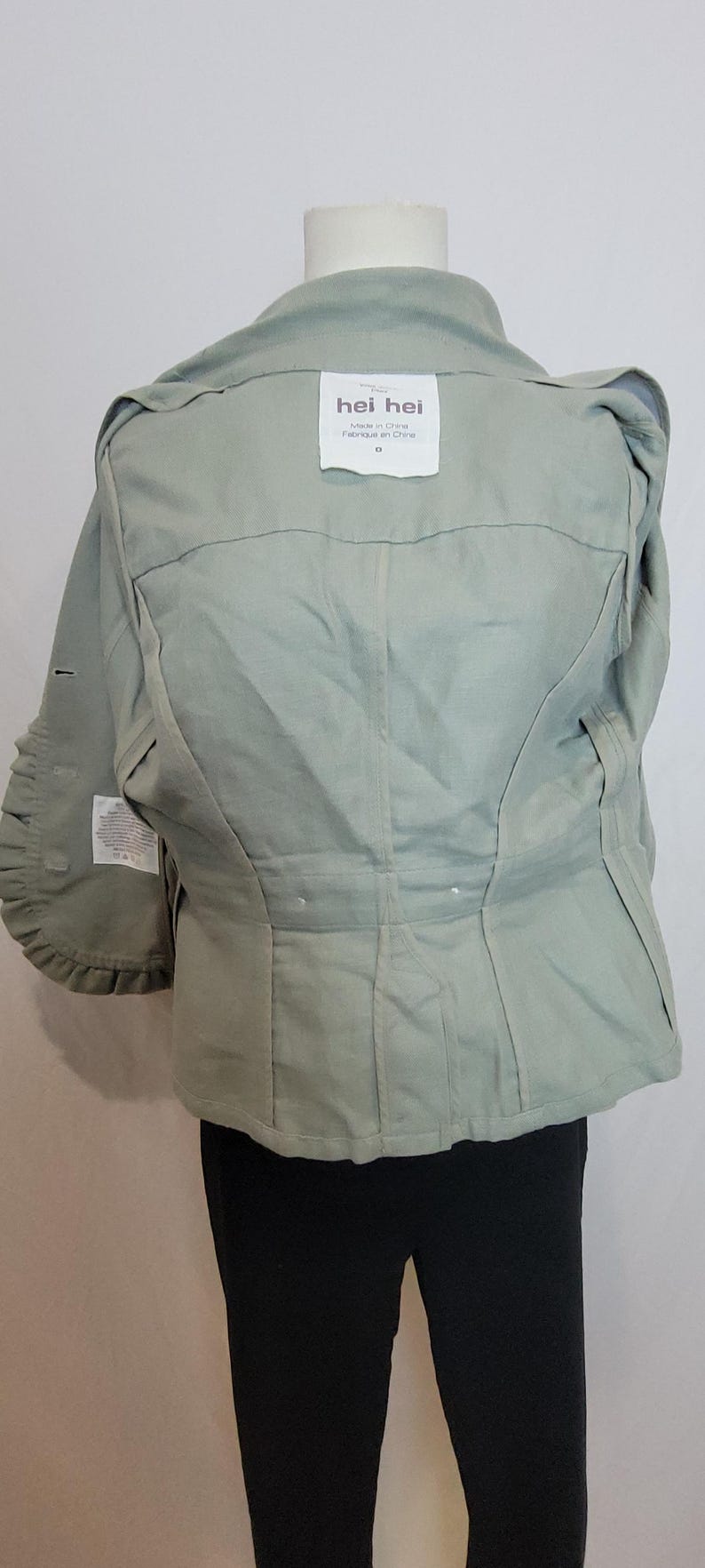 May include: A pale green jacket with a structured design. The jacket has a collar, pleats, and a label that reads "hei hei". The sleeves have a unique ruffled detail. The jacket is worn over black trousers.
