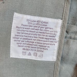 May include: A close-up of a white care label sewn into a light green garment. The label provides washing instructions in English and French, detailing washing, bleaching, drying, and ironing. The fabric is 55% linen and 45% cotton.