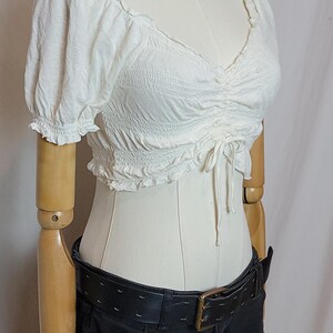 Op de afbeelding: Een witte, getextureerde crop top met korte, gerimpelde mouwen en een V-hals. De top wordt gecombineerd met een zwarte jeans en een brede zwarte riem met een gouden gesp. Een zilveren ketting is ook zichtbaar.