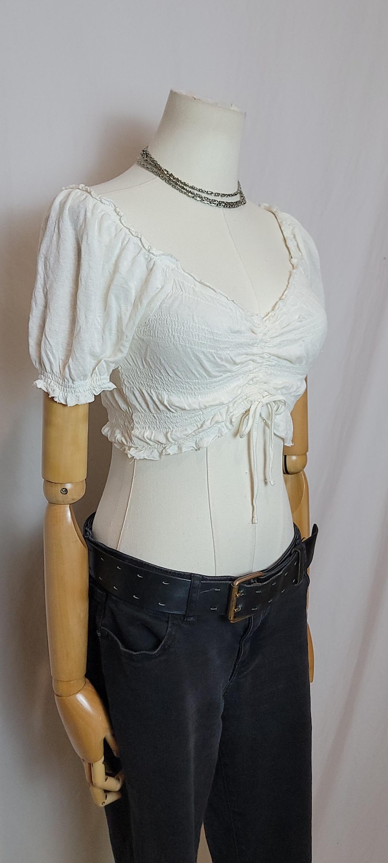 Op de afbeelding: Een witte, getextureerde crop top met korte, gerimpelde mouwen en een V-hals. De top wordt gecombineerd met een zwarte jeans en een brede zwarte riem met een gouden gesp. Een zilveren ketting is ook zichtbaar.