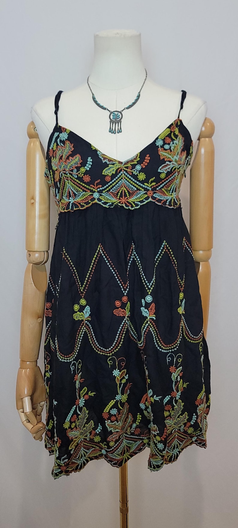 May include: Black spaghetti strap dress with colourful embroidered floral and geometric patterns. The dress features a V-neckline and a fitted bodice, displayed on a mannequin. A silver necklace with a pendant is also visible.