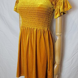 May include: Mustard yellow velvet dress with a square neckline and short, ruffled sleeves. The bodice is smocked, and the skirt is flared. A multi-colored beaded necklace is around the mannequin's neck. The dress is displayed on a wooden mannequin.